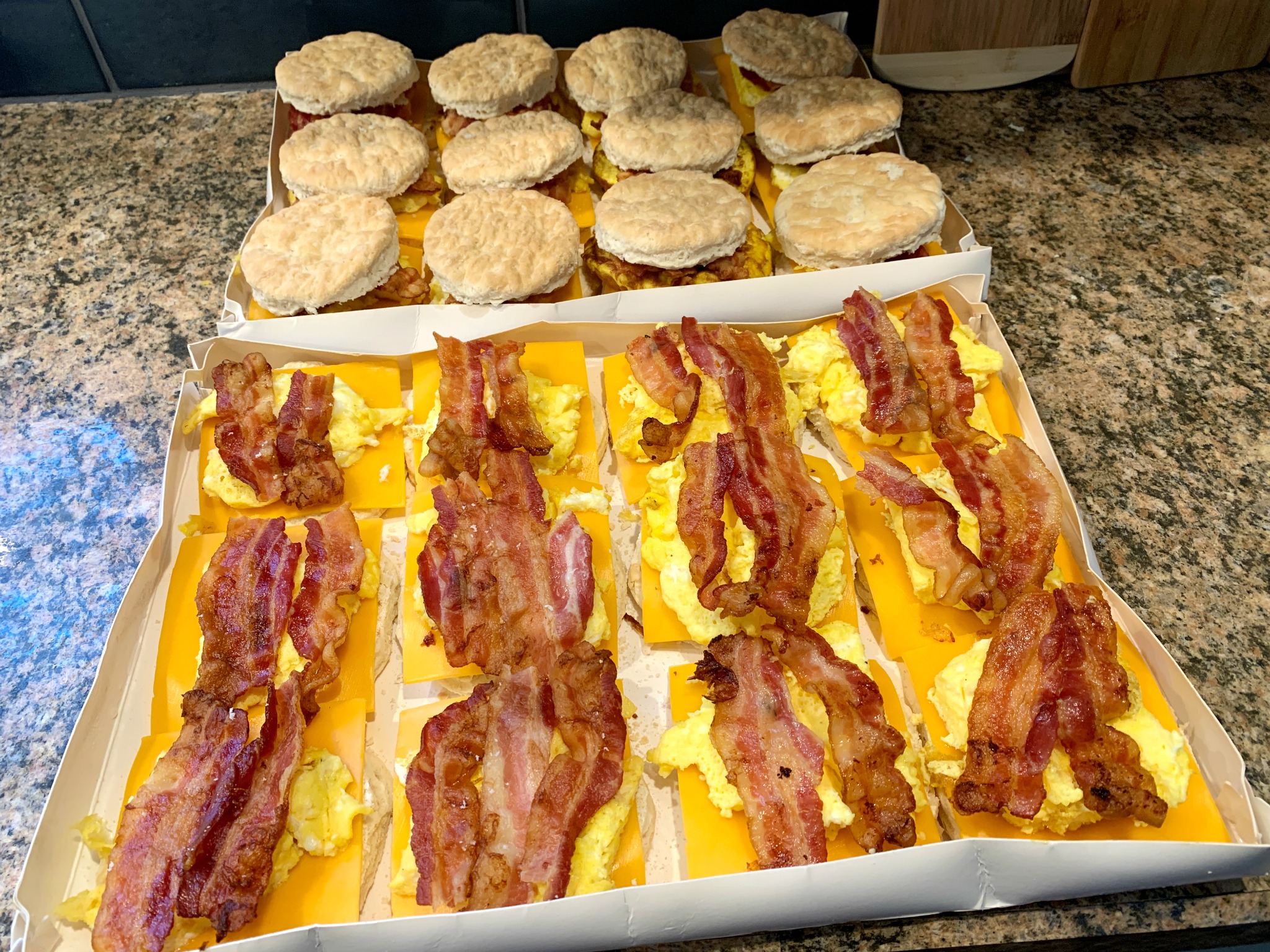 a dozen bacon, egg and cheese sandwiches without the top with another dozen sandwiches in the background with the top biscuit on