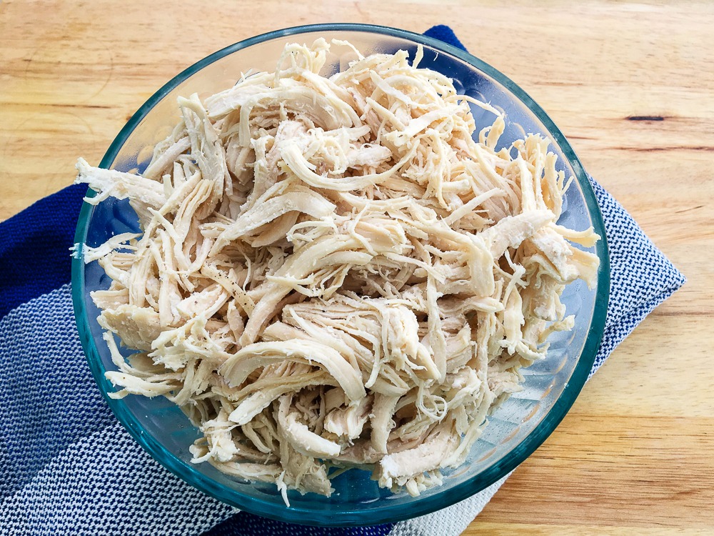 a glass bowl filled with shredded chicken with a blue tea towel under it