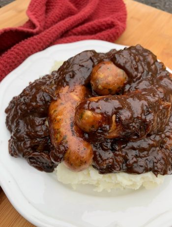 a white plate with mashed potatoes, sausages and onion gravy poured on top
