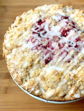 round coffee cake with crumb topping, raspberries in the center and a powdered sugar glaze