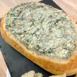 an oval loaf of bread filed with spinach dip on a piece of slate