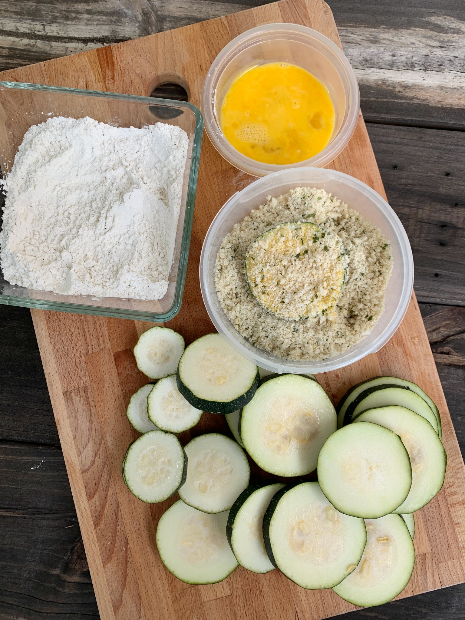 wooden cutting board with sliced zucchini, a bowl of panko and parmesan mixed, a bowl of flour and a bowl of beaten eggs