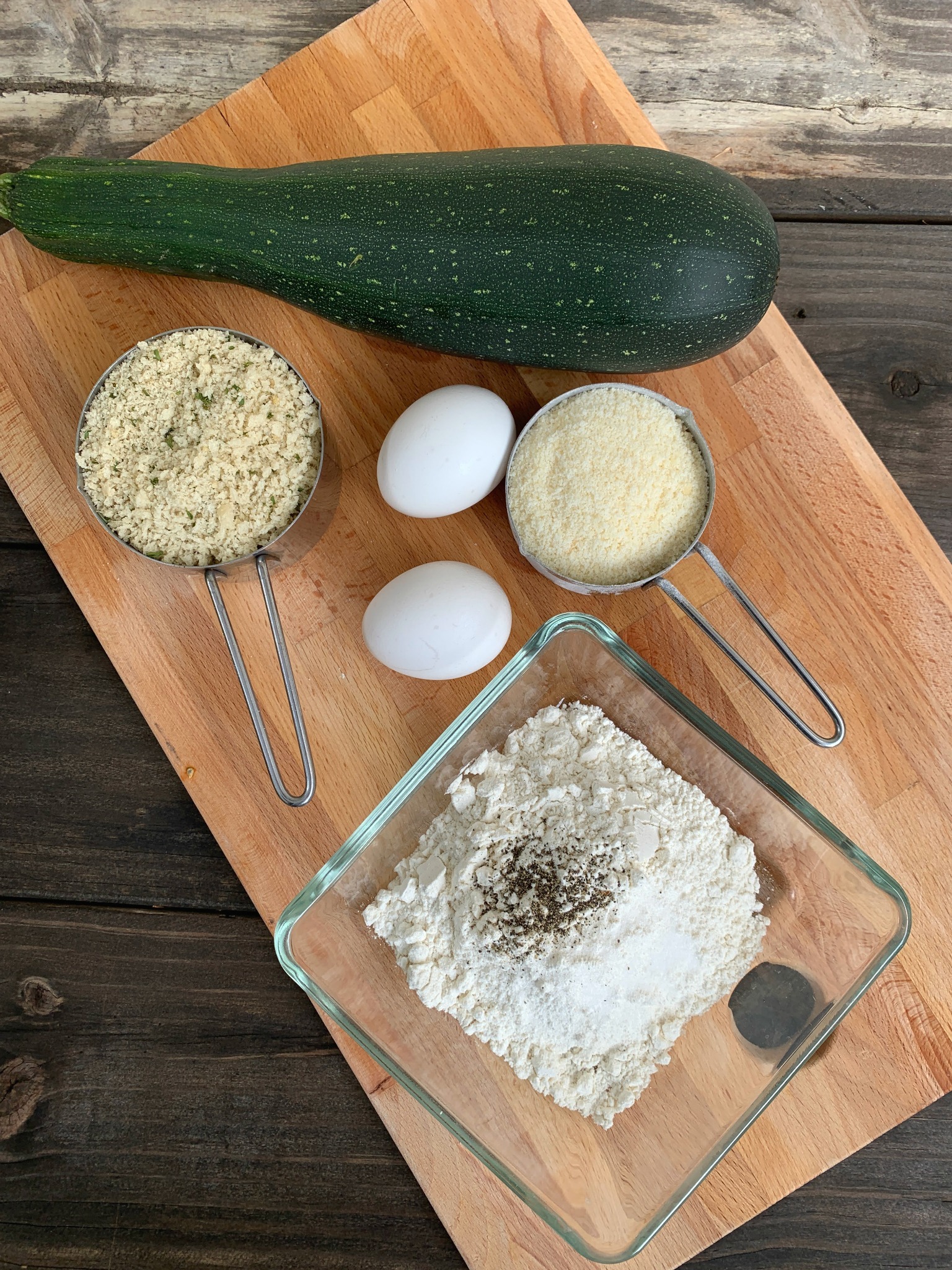 wooden cutting board with zucchini, measuring cups with panko bread crumbs and parmesan, 2 eggs and a glass bowl with flour